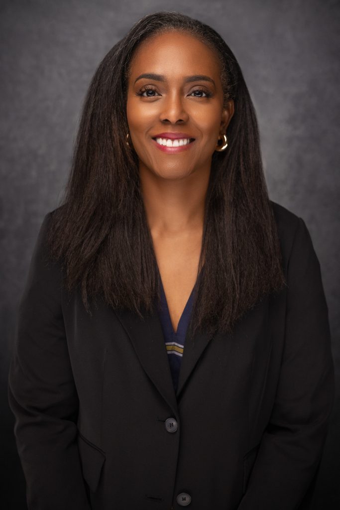A woman with long, straight dark hair smiles at the camera. She is wearing a black blazer over a dark top and gold hoop earrings, standing against a dark gray background.