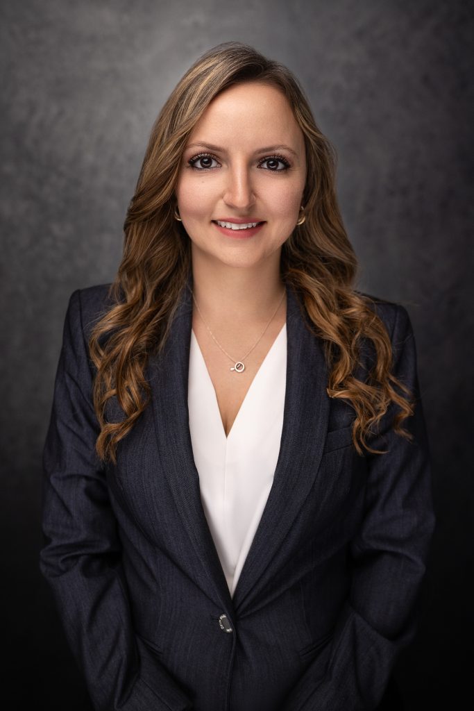 A woman with long, wavy brown hair, wearing a dark blazer and white blouse, smiles at the camera against a dark, textured background.