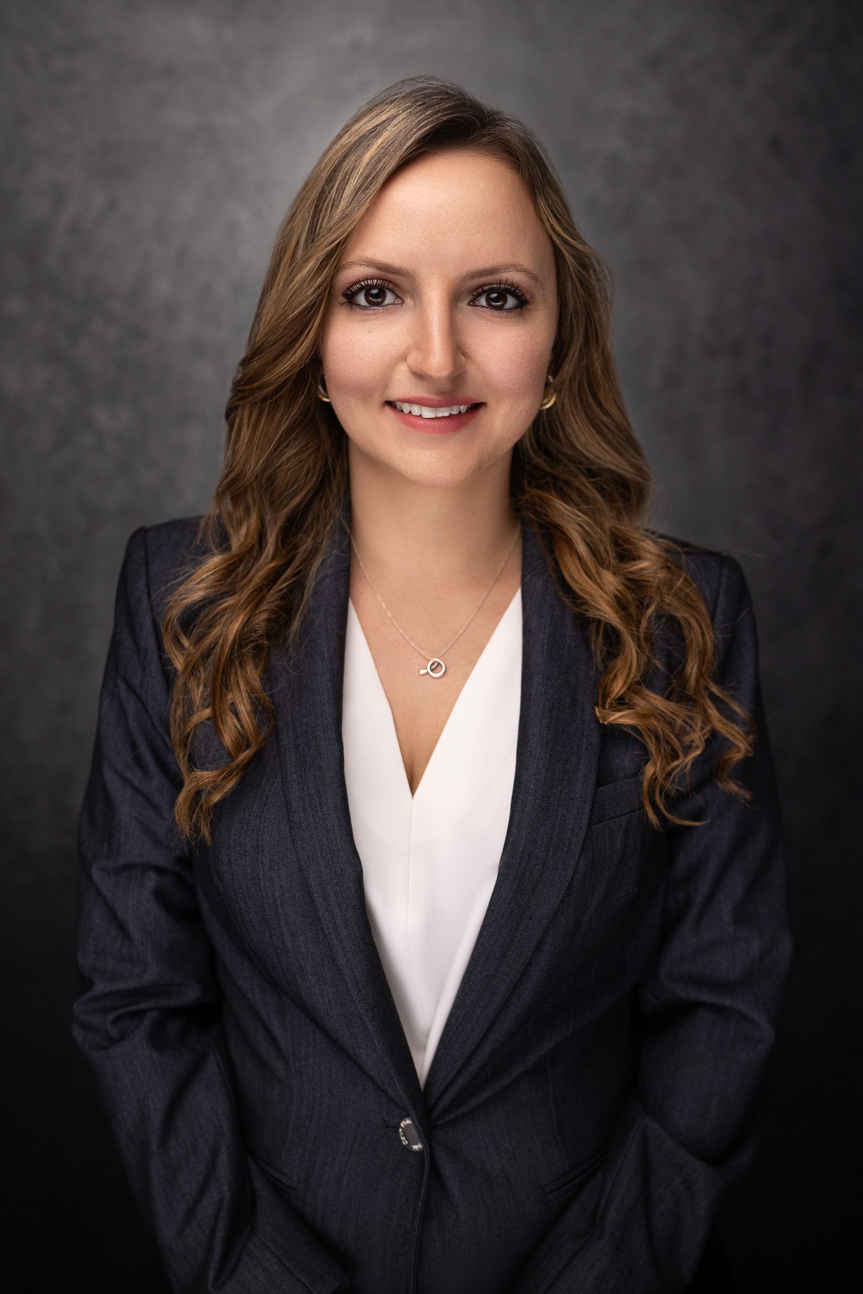 A woman with long, wavy brown hair, wearing a dark blazer and white blouse, smiles at the camera against a dark, textured background.