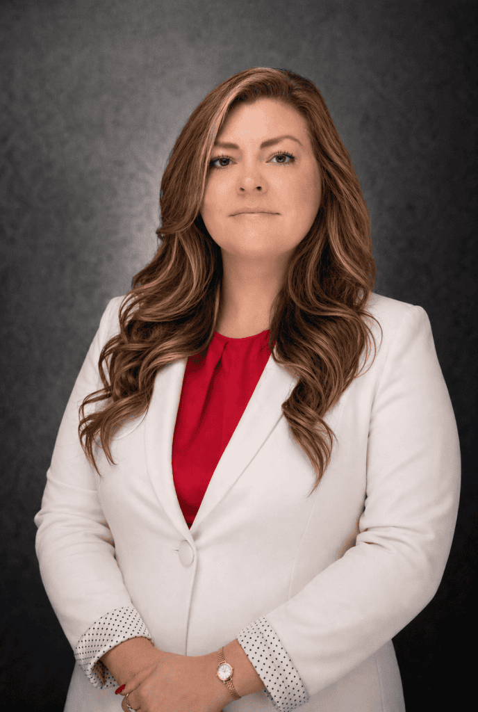 A woman with long, wavy brown hair wearing a white blazer over a red top stands in front of a dark, textured background, looking confidently at the camera with her hands clasped in front of her.