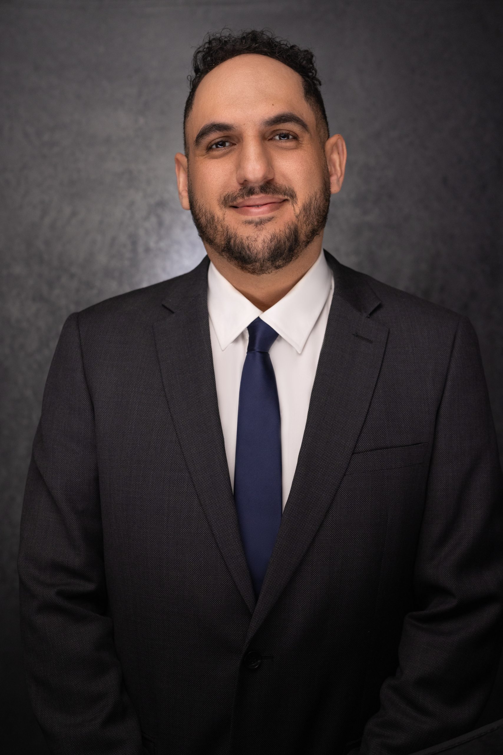 A man with short, dark curly hair and a trimmed beard, wearing a dark suit, white shirt, and navy tie, stands smiling in front of a dark, textured background.