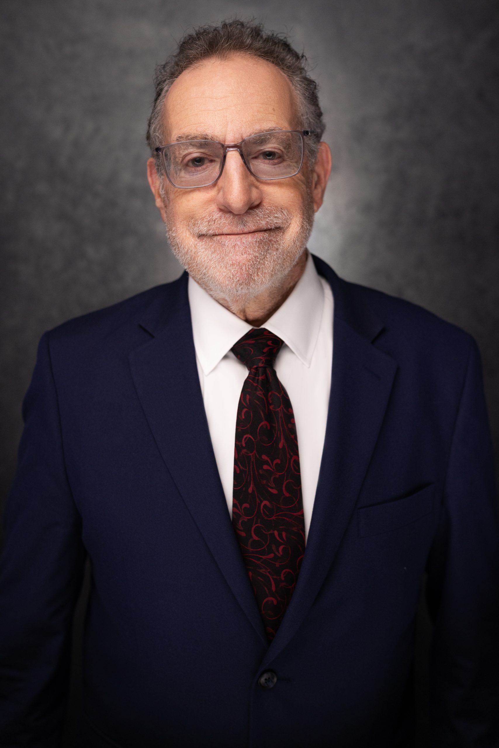 An older man with gray hair, glasses, and a trimmed beard smiles softly. He is wearing a navy blue suit, white shirt, and a dark tie with a red pattern, standing against a dark, textured background.