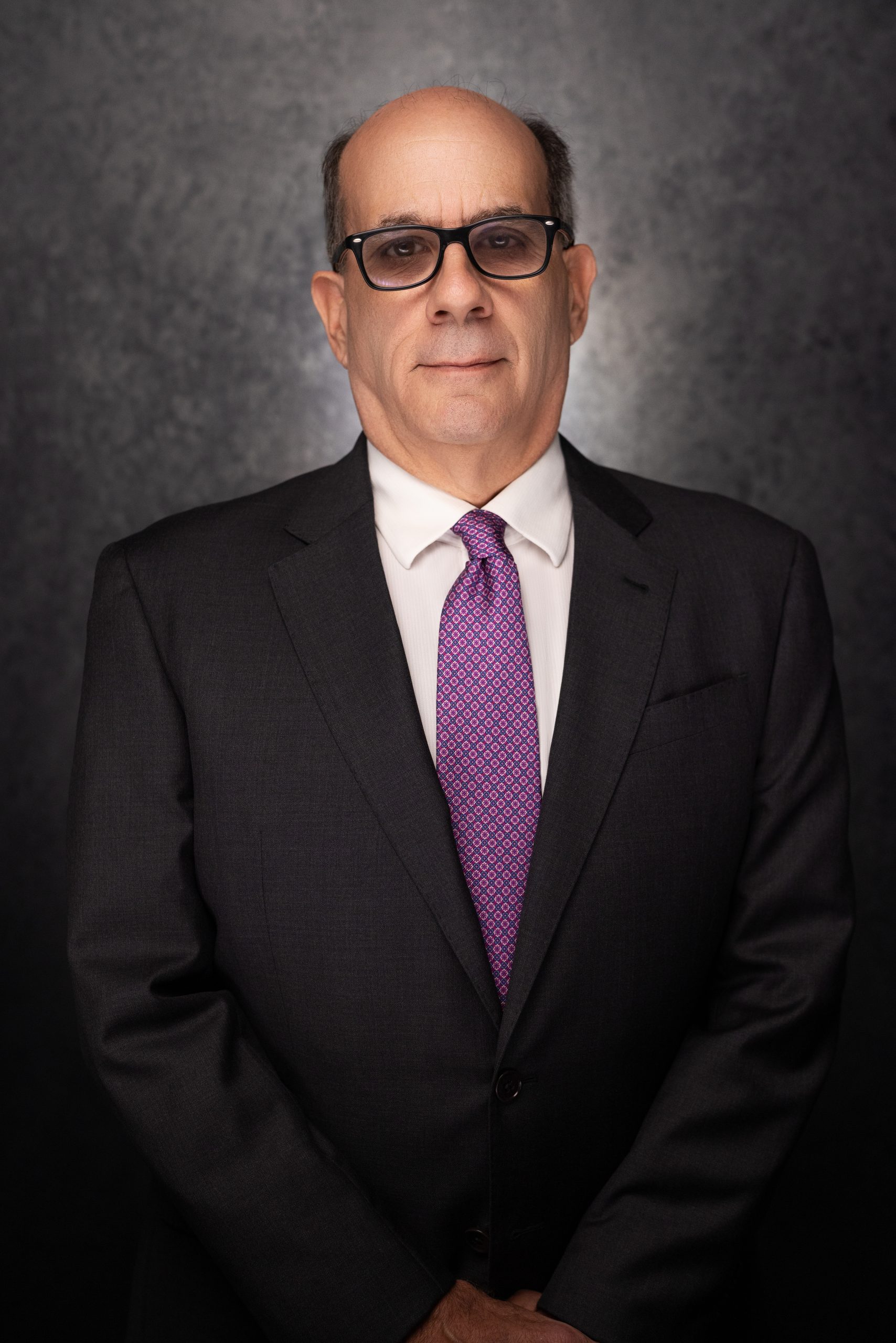 A man in a dark suit, white shirt, and purple patterned tie stands against a dark, textured background. He has short hair, wears glasses, and faces the camera with a neutral expression.