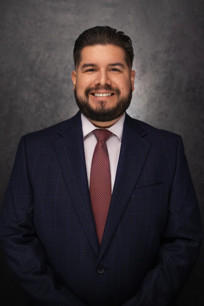 A man with dark hair and a beard, wearing a blue checkered suit, white shirt, and maroon tie, smiles while standing against a dark, textured background.