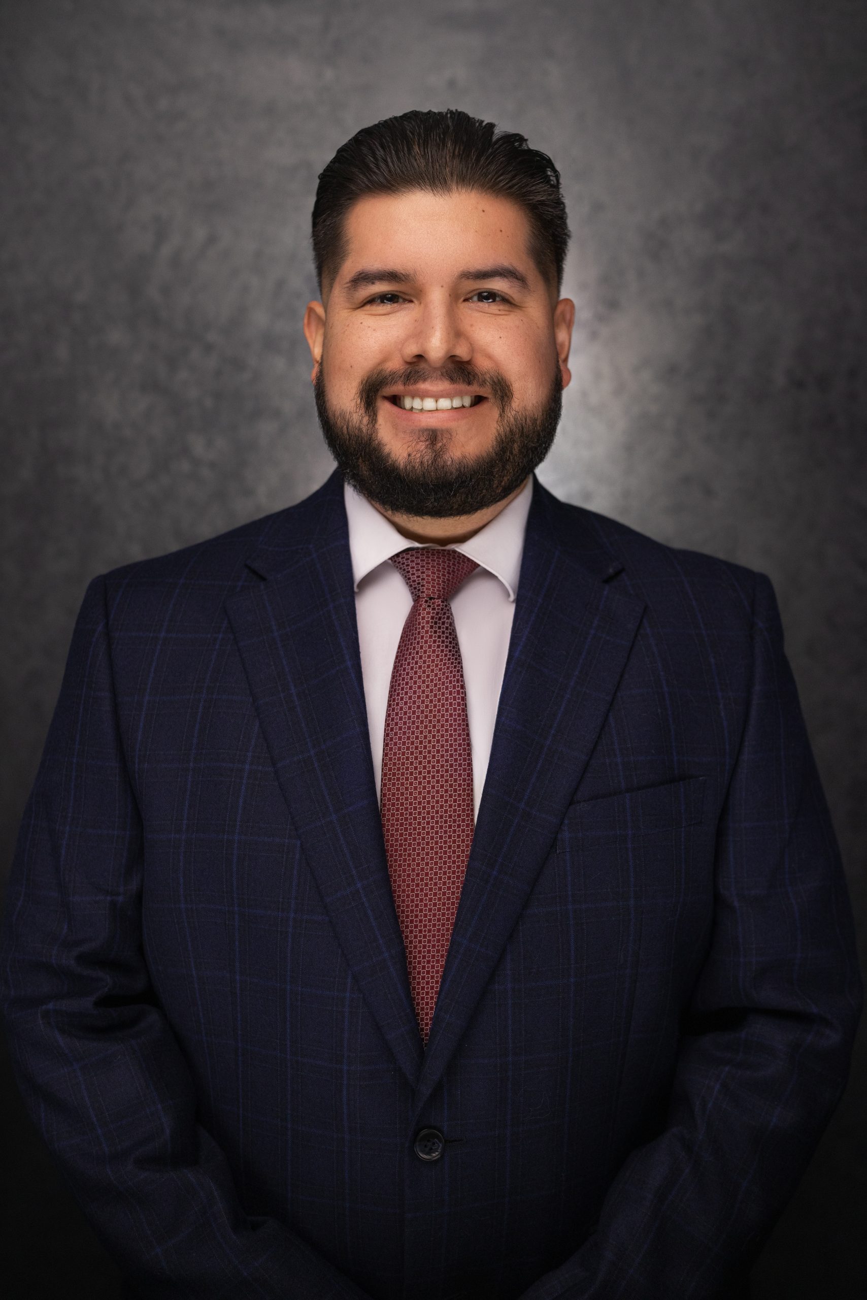 A man with dark hair and a beard, wearing a blue checkered suit, white shirt, and maroon tie, smiles while standing against a dark, textured background.