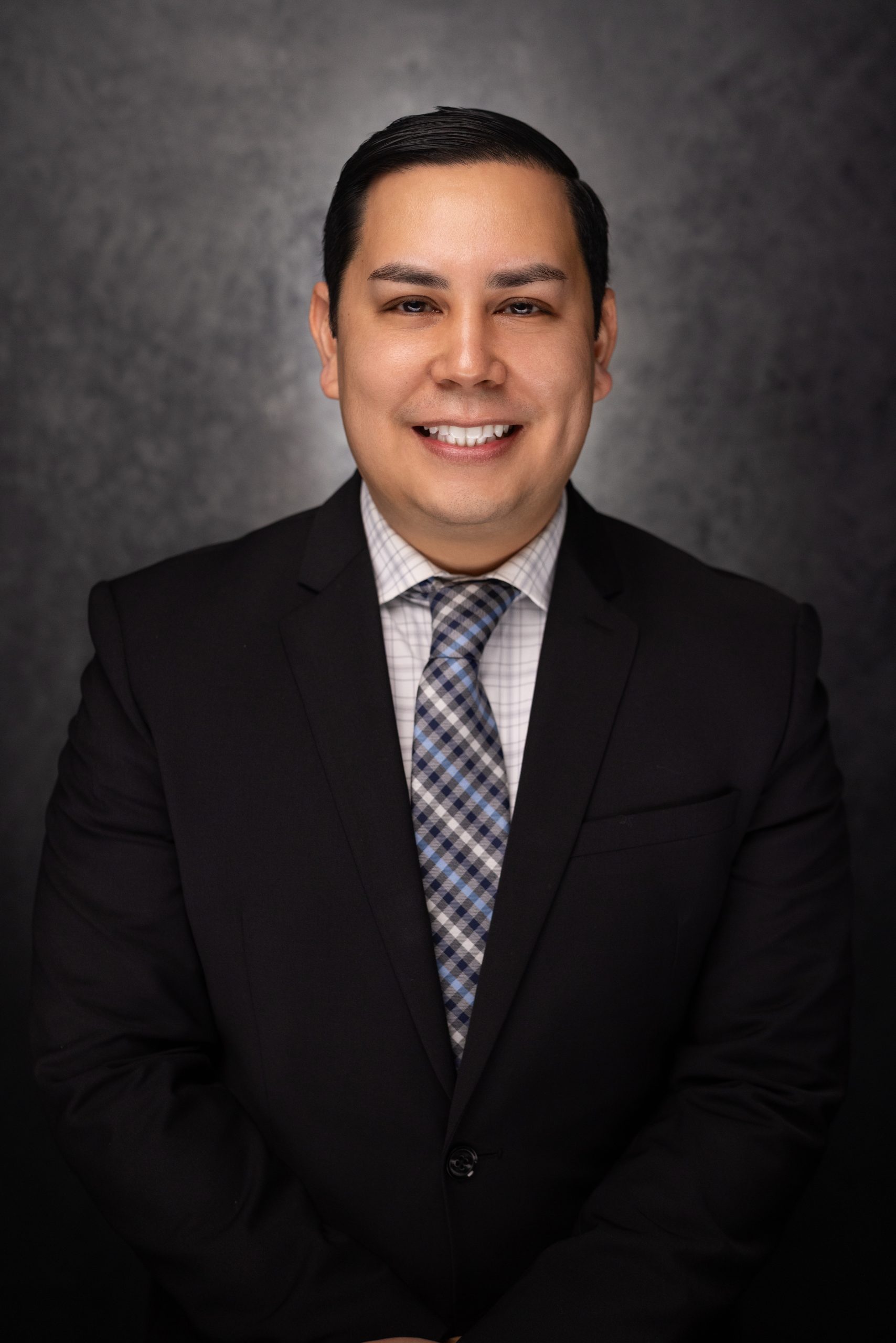 A man in a dark suit, patterned tie, and checked shirt smiles at the camera against a dark, textured background. He has short, dark hair and is posing for a professional portrait.