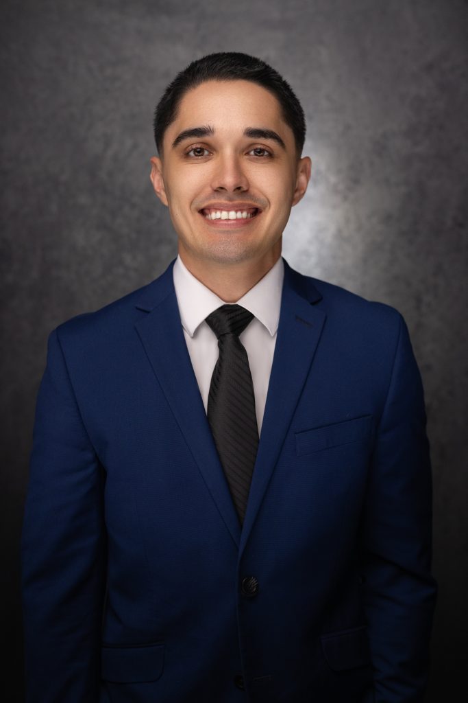 A young man in a blue suit, white dress shirt, and black tie smiles at the camera in front of a dark, textured background.