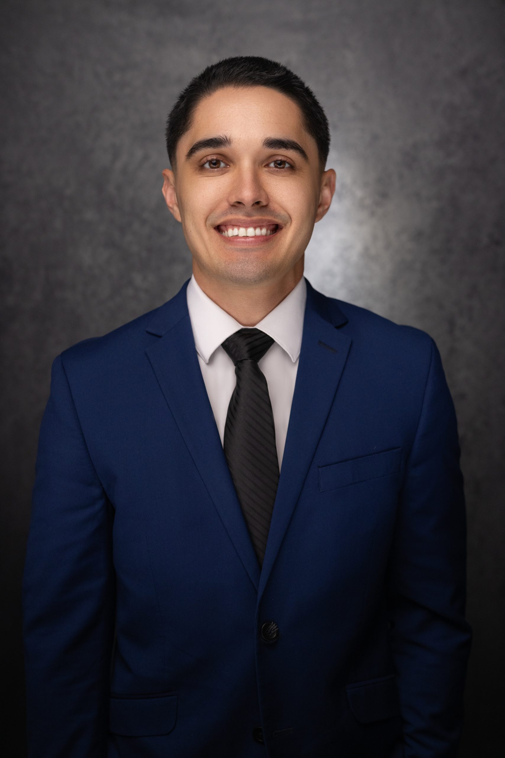 A young man in a blue suit, white dress shirt, and black tie smiles at the camera in front of a dark, textured background.