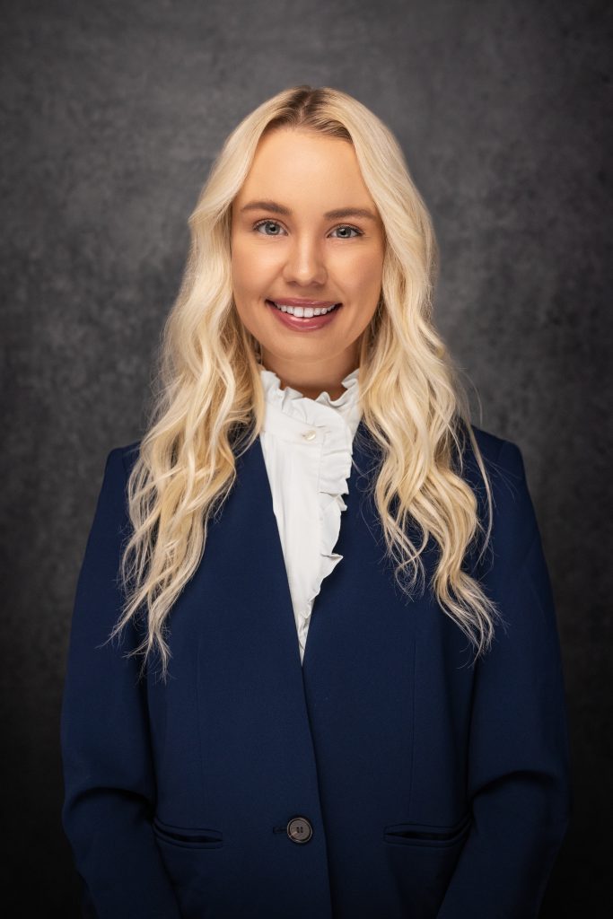 A woman with long, wavy blonde hair, wearing a navy blue blazer and a white ruffled blouse, smiles in front of a dark, textured background.