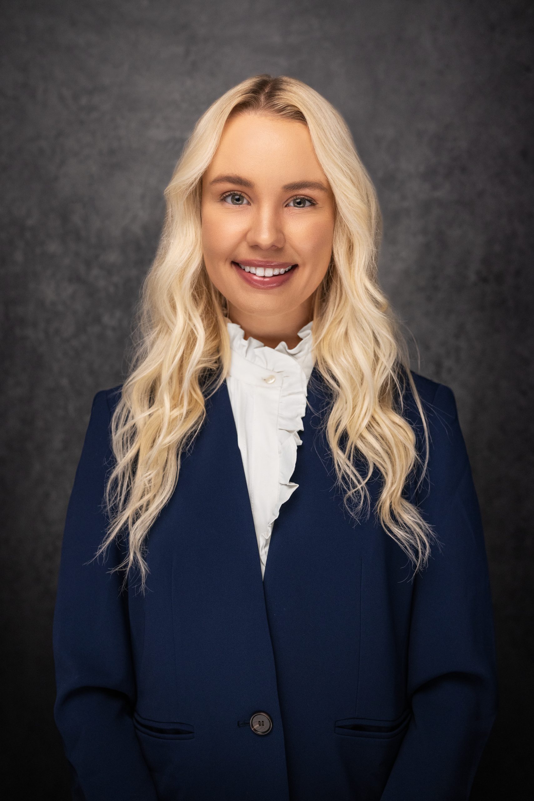 A woman with long, wavy blonde hair, wearing a navy blue blazer and a white ruffled blouse, smiles in front of a dark, textured background.