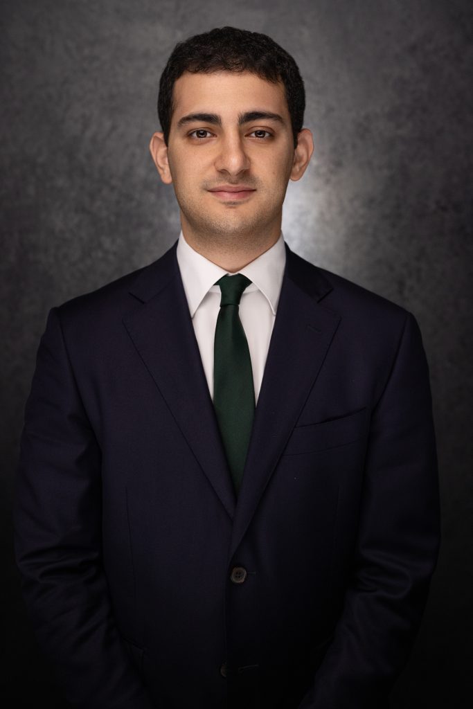 A man with short dark hair wearing a navy suit, white shirt, and dark green tie stands in front of a dark, textured background, looking at the camera with a neutral expression.