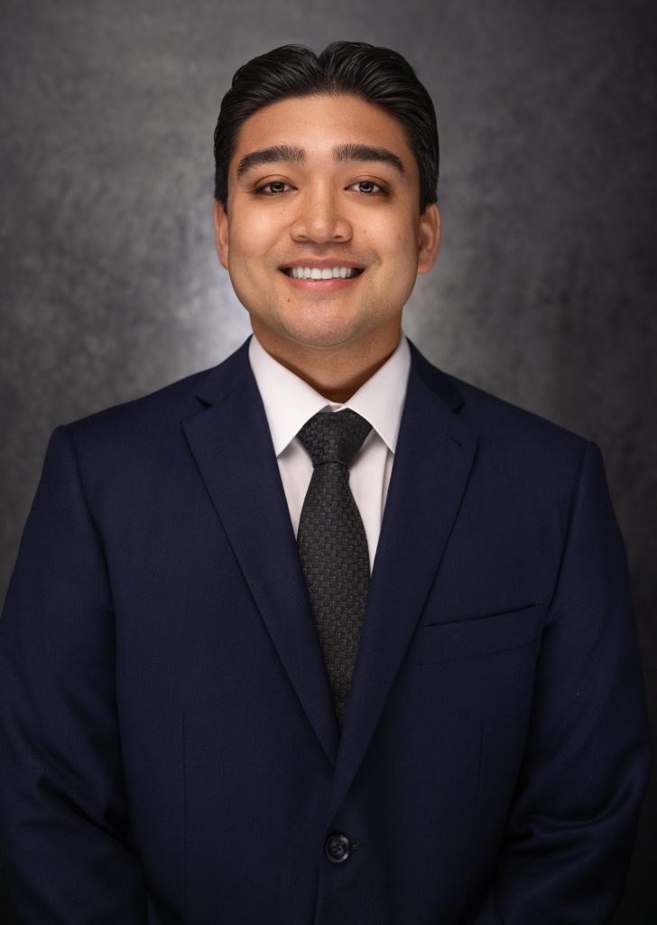 A man wearing a dark blue suit, white shirt, and dark tie smiles at the camera. He has short dark hair and stands against a dark, textured background.