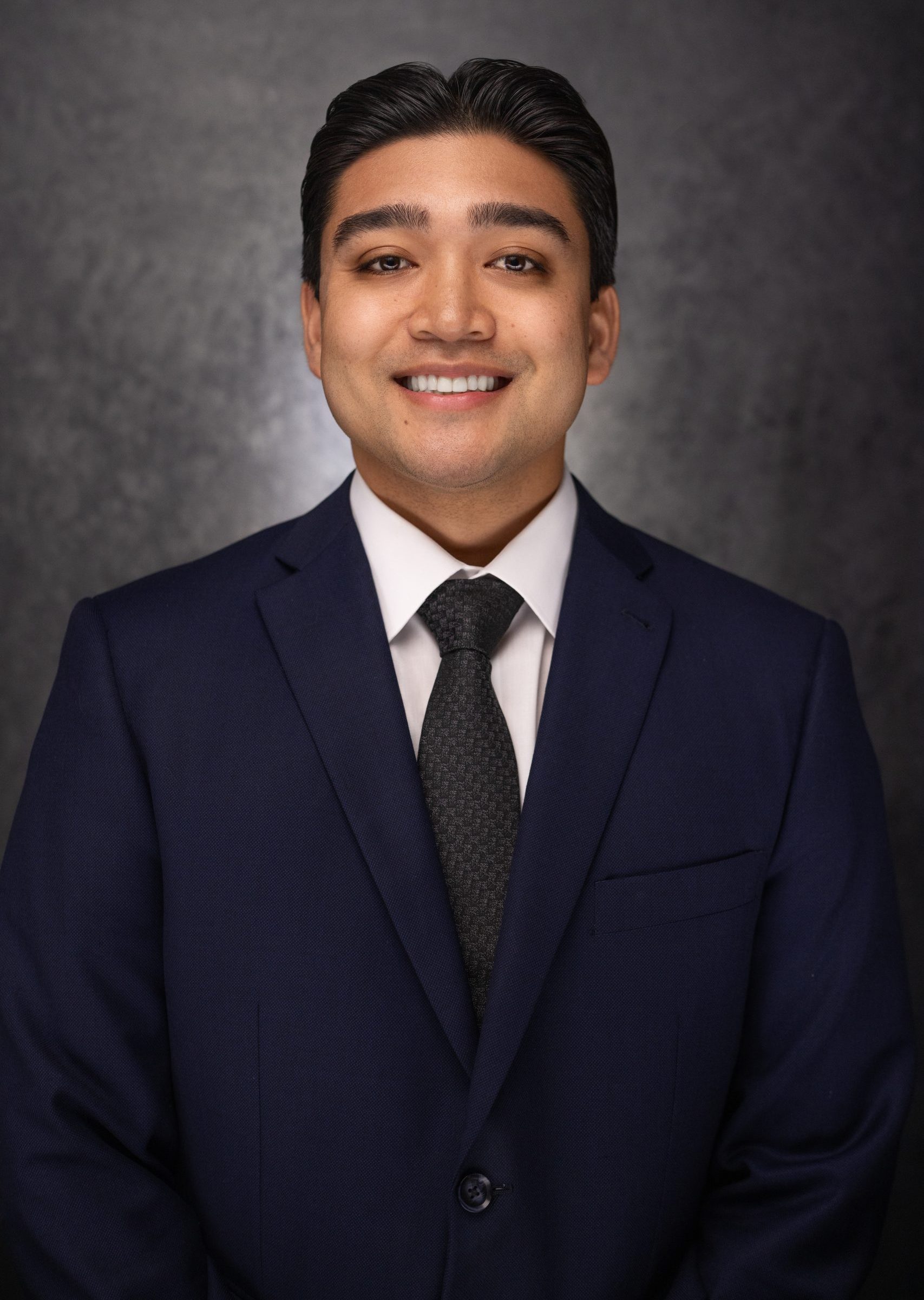 A man wearing a dark blue suit, white shirt, and dark tie smiles at the camera. He has short dark hair and stands against a dark, textured background.