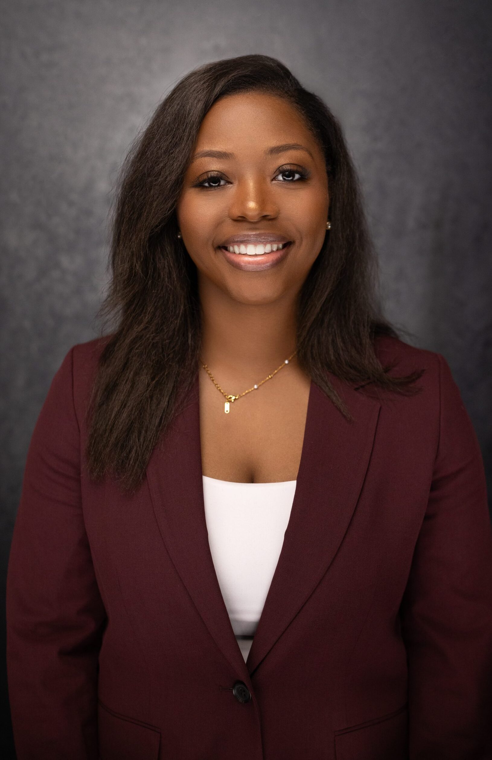 A woman wearing a maroon blazer over a white top smiles at the camera against a dark, textured background. She has straight, shoulder-length hair and wears a gold necklace.