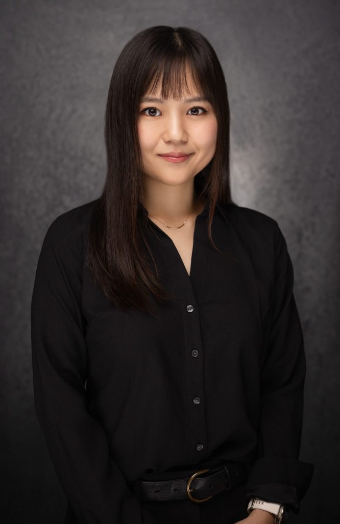 A young woman with long, straight dark hair stands in front of a dark, textured background. She is wearing a black button-up shirt and has a slight smile, looking directly at the camera.