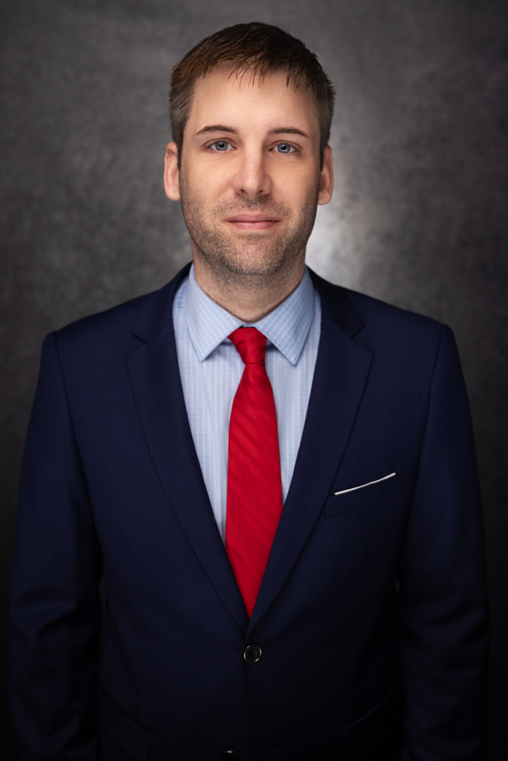 A man wearing a navy blue suit, light blue shirt, and red tie stands in front of a dark, textured background, looking directly at the camera with a neutral expression.