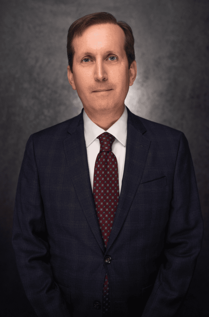 A middle-aged man with short brown hair, wearing a dark blue suit, white dress shirt, and patterned red tie, poses against a dark, textured background. He is facing the camera with a neutral expression.