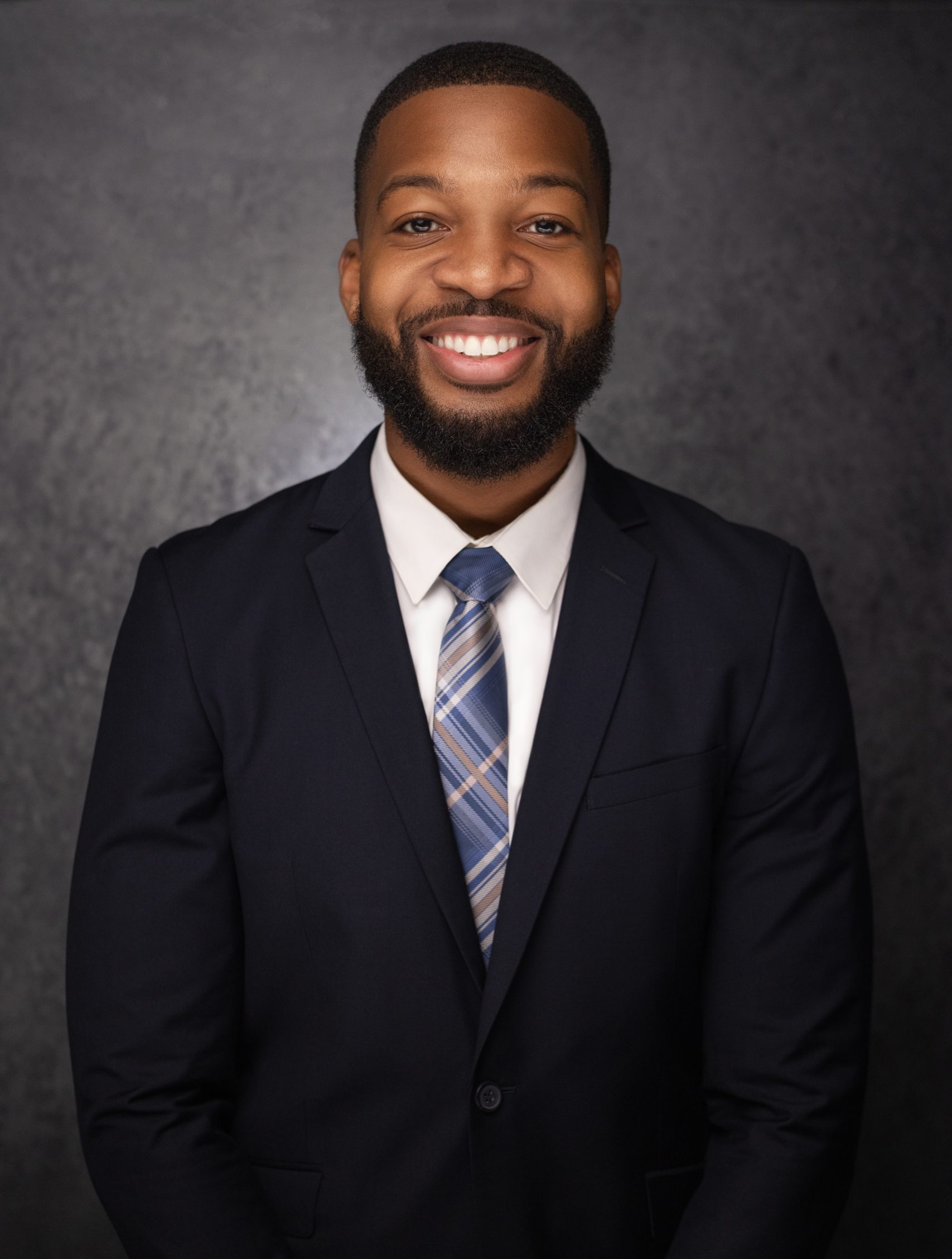 A smiling man in a dark suit, white shirt, and striped tie stands against a dark, textured background, looking confidently at the camera.