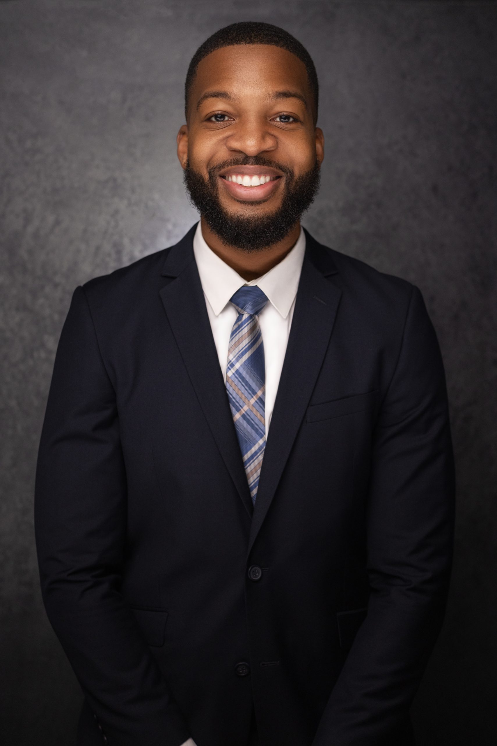 A smiling man in a dark suit, white shirt, and striped tie stands against a dark, textured background, looking confidently at the camera.