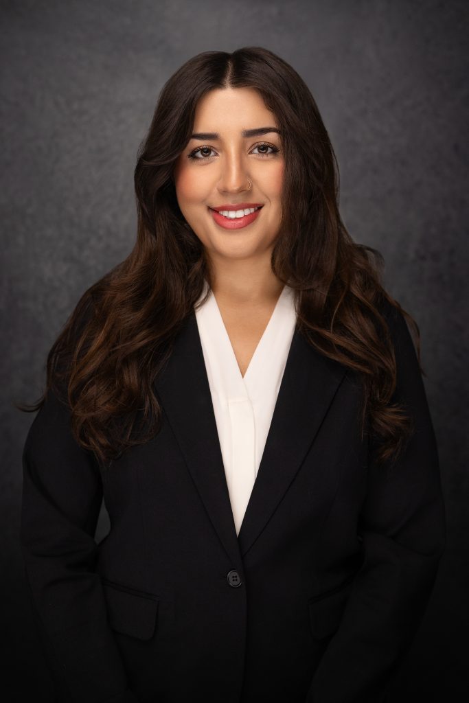 A woman with long brown hair wearing a black blazer and white blouse smiles at the camera in front of a dark, textured background.
