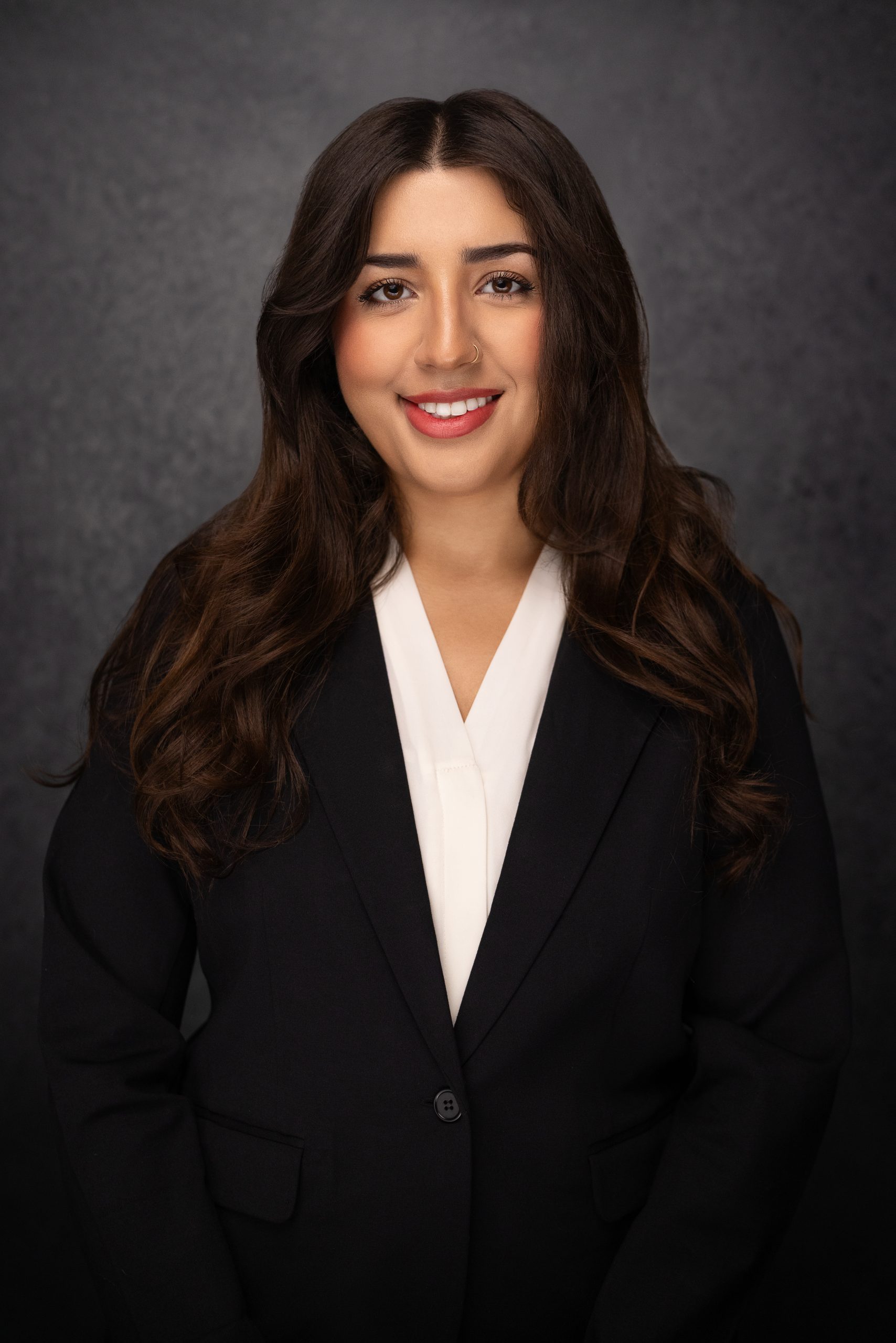 A woman with long brown hair wearing a black blazer and white blouse smiles at the camera in front of a dark, textured background.