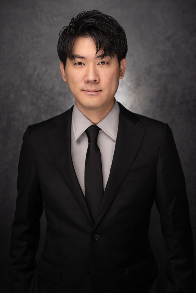 A man with short black hair wears a black suit, black tie, and light gray shirt, standing in front of a dark, textured background, facing the camera with a neutral expression.