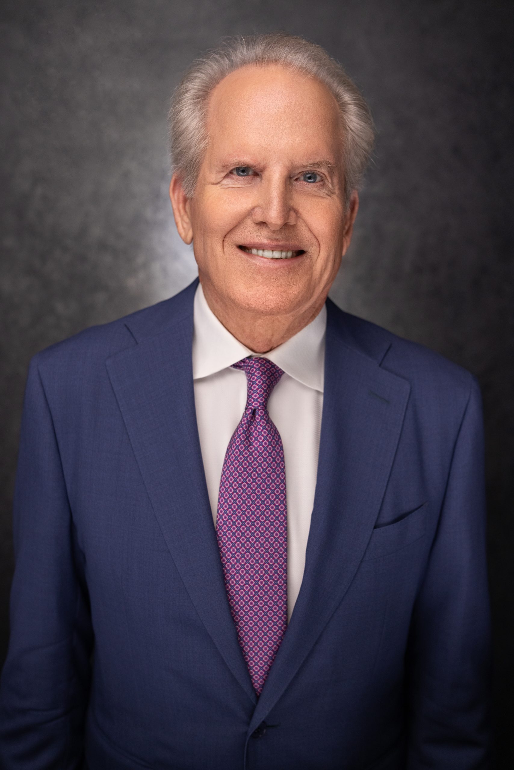 A smiling older man with gray hair wearing a blue suit, white shirt, and patterned purple tie, stands in front of a dark, textured background.