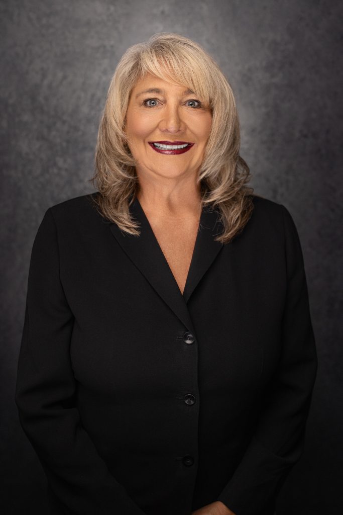 A woman with blonde hair wearing a black blazer smiles in front of a dark, textured background.