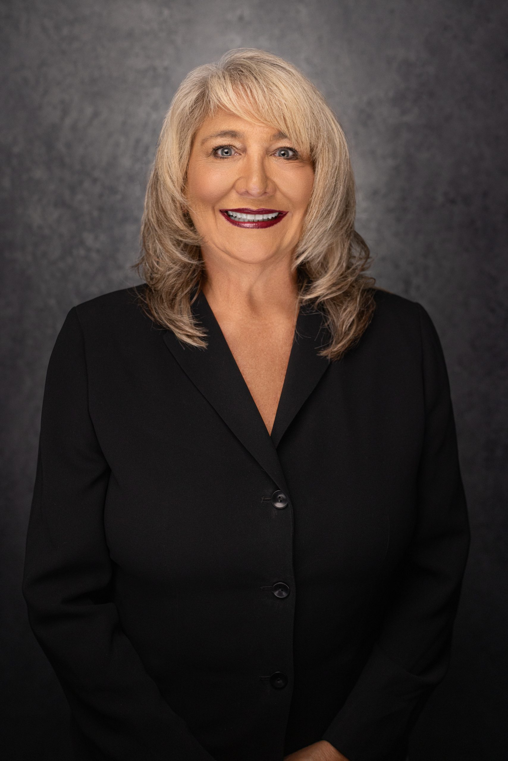 A woman with blonde hair wearing a black blazer smiles in front of a dark, textured background.