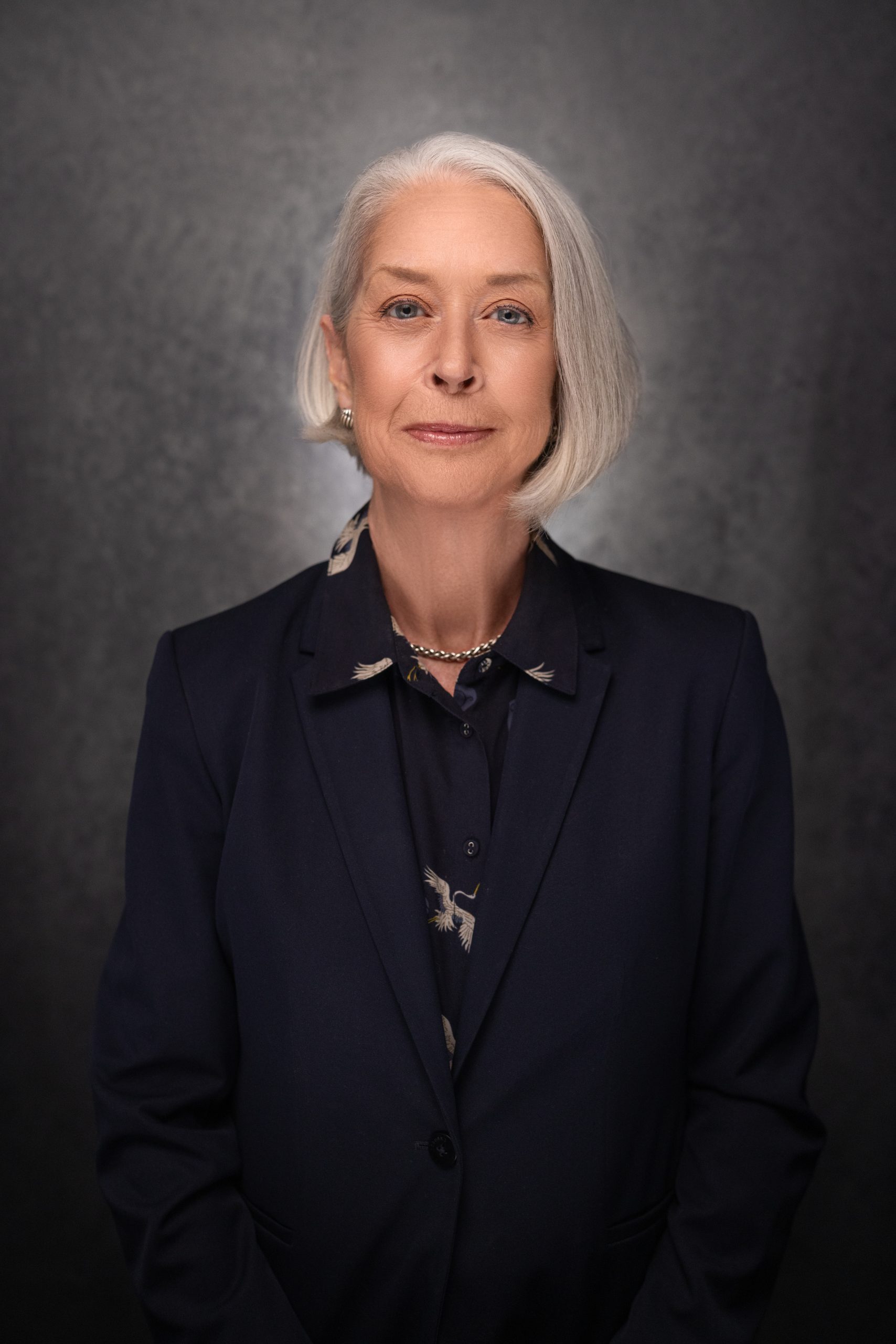 Portrait of an older woman with short gray hair wearing a dark navy blazer and patterned blouse, standing against a dark gray textured background, looking confidently at the camera.