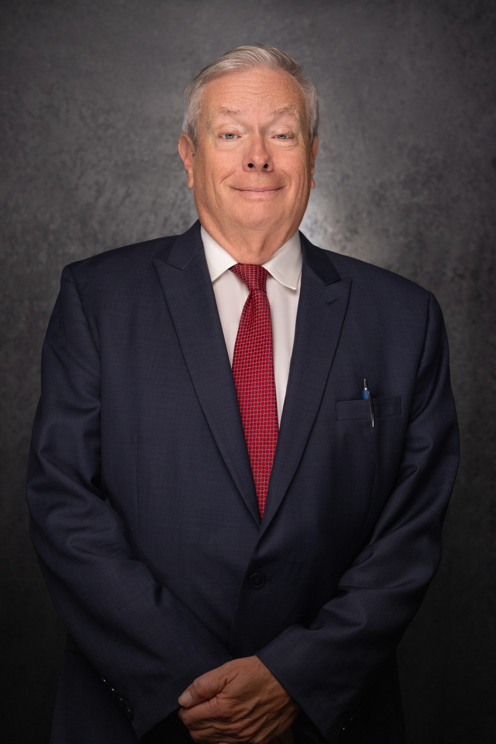 An older man in a navy suit, white shirt, and red tie stands in front of a dark, textured background, smiling slightly with his hands clasped in front of him. A pen is visible in his jacket pocket.