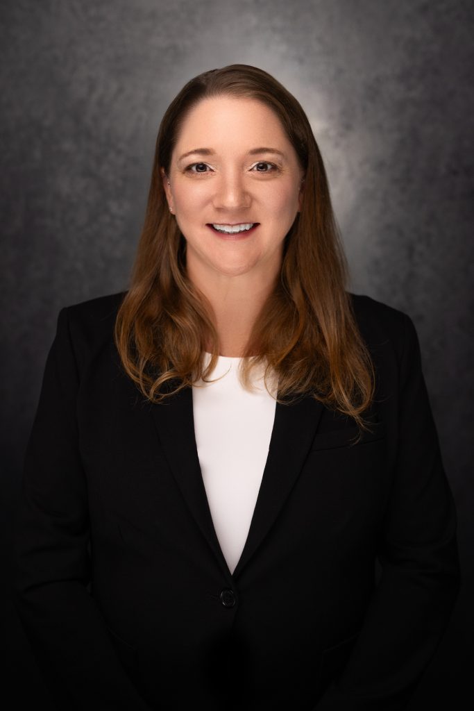 A woman with long brown hair, wearing a black blazer and white top, is smiling at the camera against a dark, textured background.