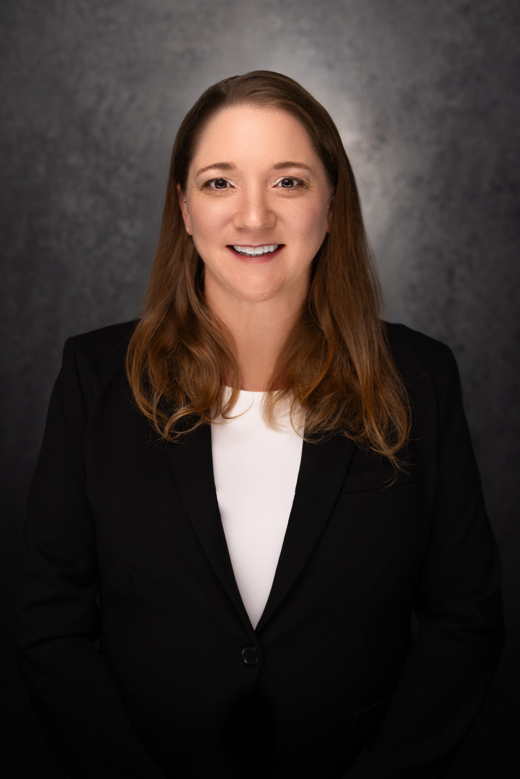 A woman with long brown hair, wearing a black blazer and white top, is smiling at the camera against a dark, textured background.