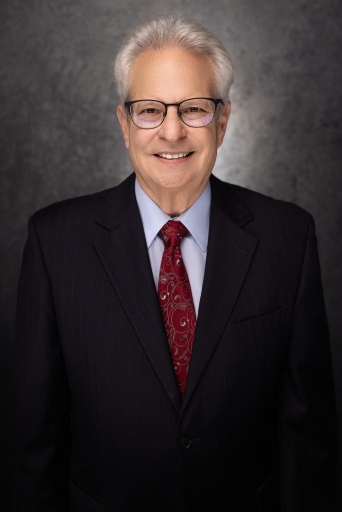 Smiling older man with gray hair and glasses, wearing a dark pinstripe suit, light blue shirt, and red patterned tie, posing in front of a dark, textured background.