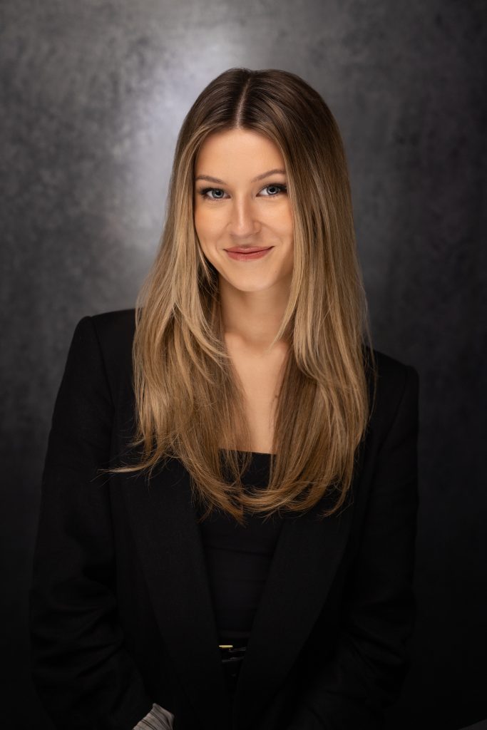 A young woman with long, straight blonde hair smiles softly at the camera. She is wearing a black blazer over a black top and is posed against a dark, textured background.