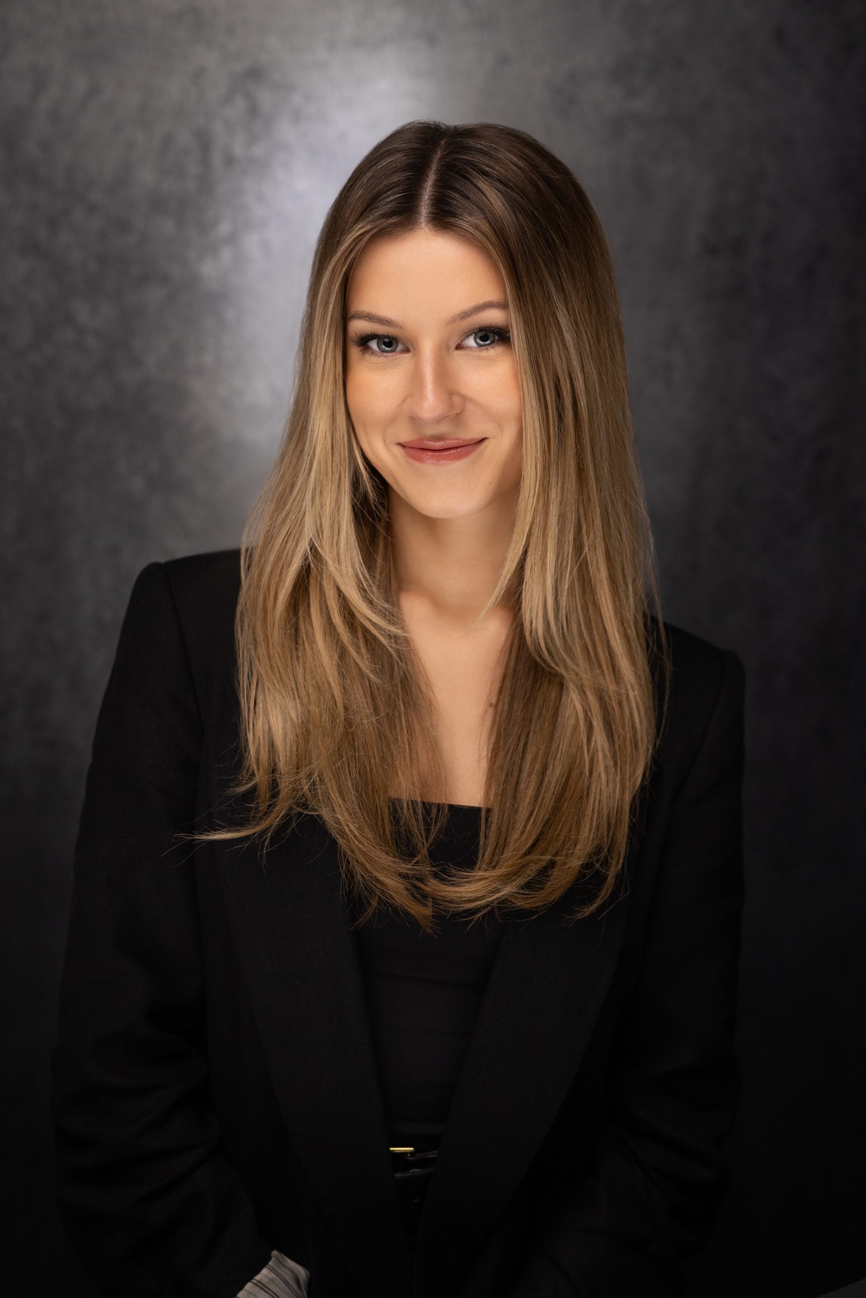 A young woman with long, straight blonde hair smiles softly at the camera. She is wearing a black blazer over a black top and is posed against a dark, textured background.