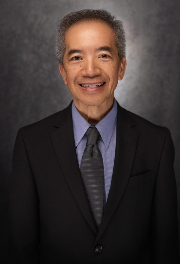 A smiling older man with short gray hair wears a black suit, blue shirt, and dark tie, standing in front of a dark, textured background.