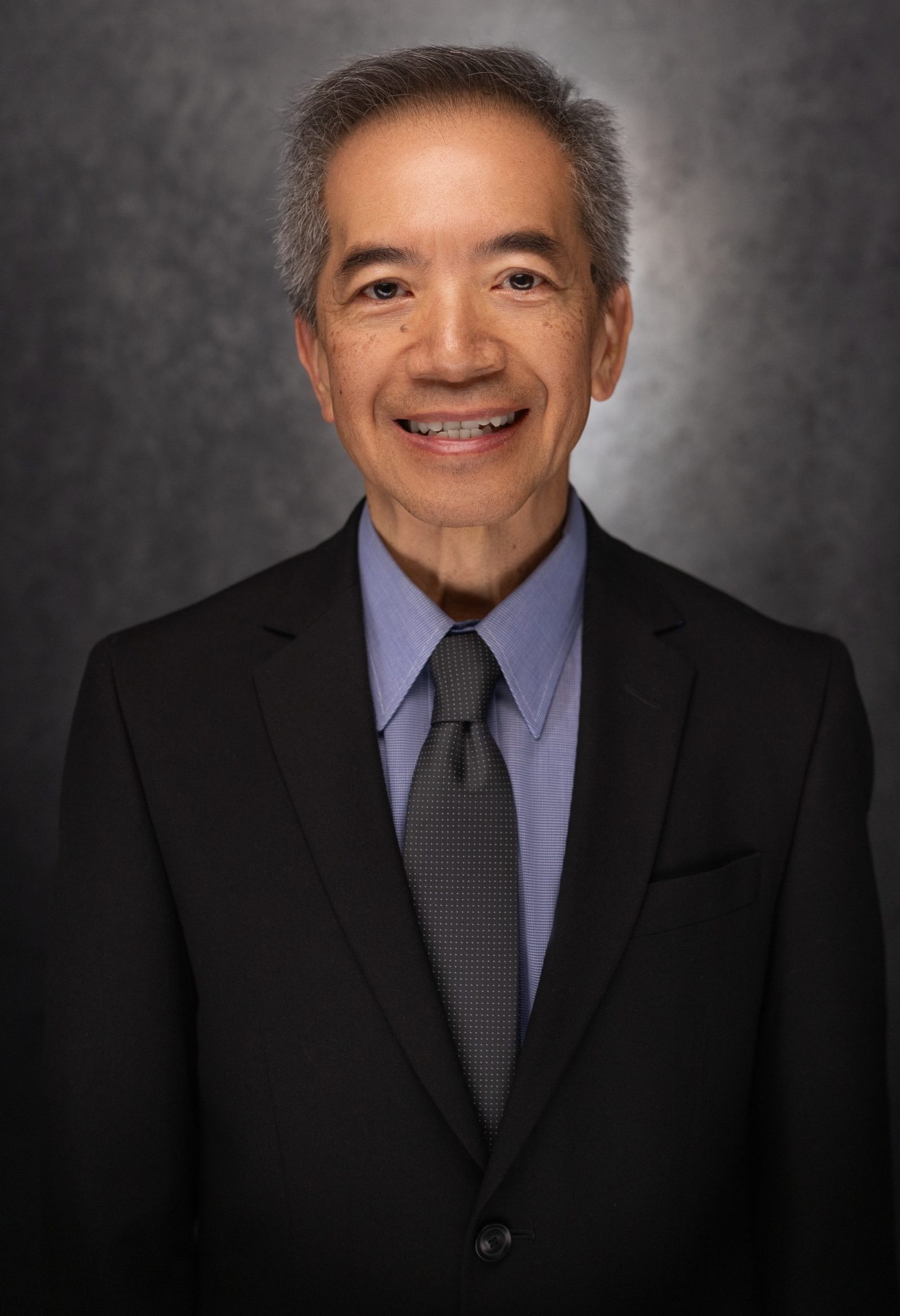 A smiling older man with short gray hair wears a black suit, blue shirt, and dark tie, standing in front of a dark, textured background.