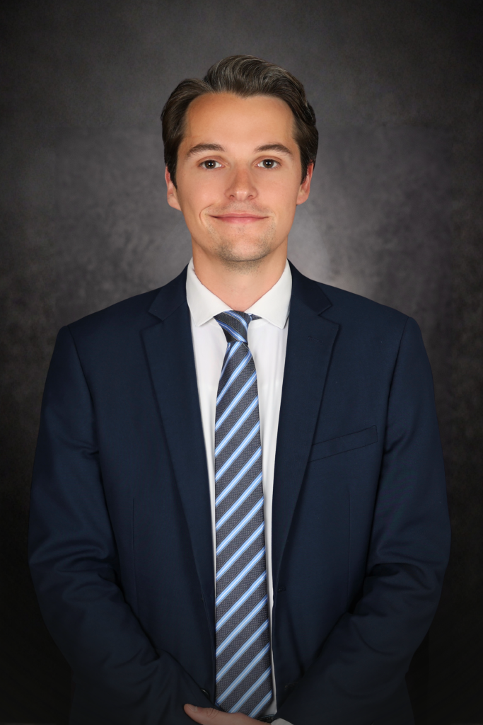 A young man with short brown hair wearing a dark blue suit, white dress shirt, and a striped blue tie stands against a dark, textured background, smiling gently at the camera with his hands clasped in front of him.