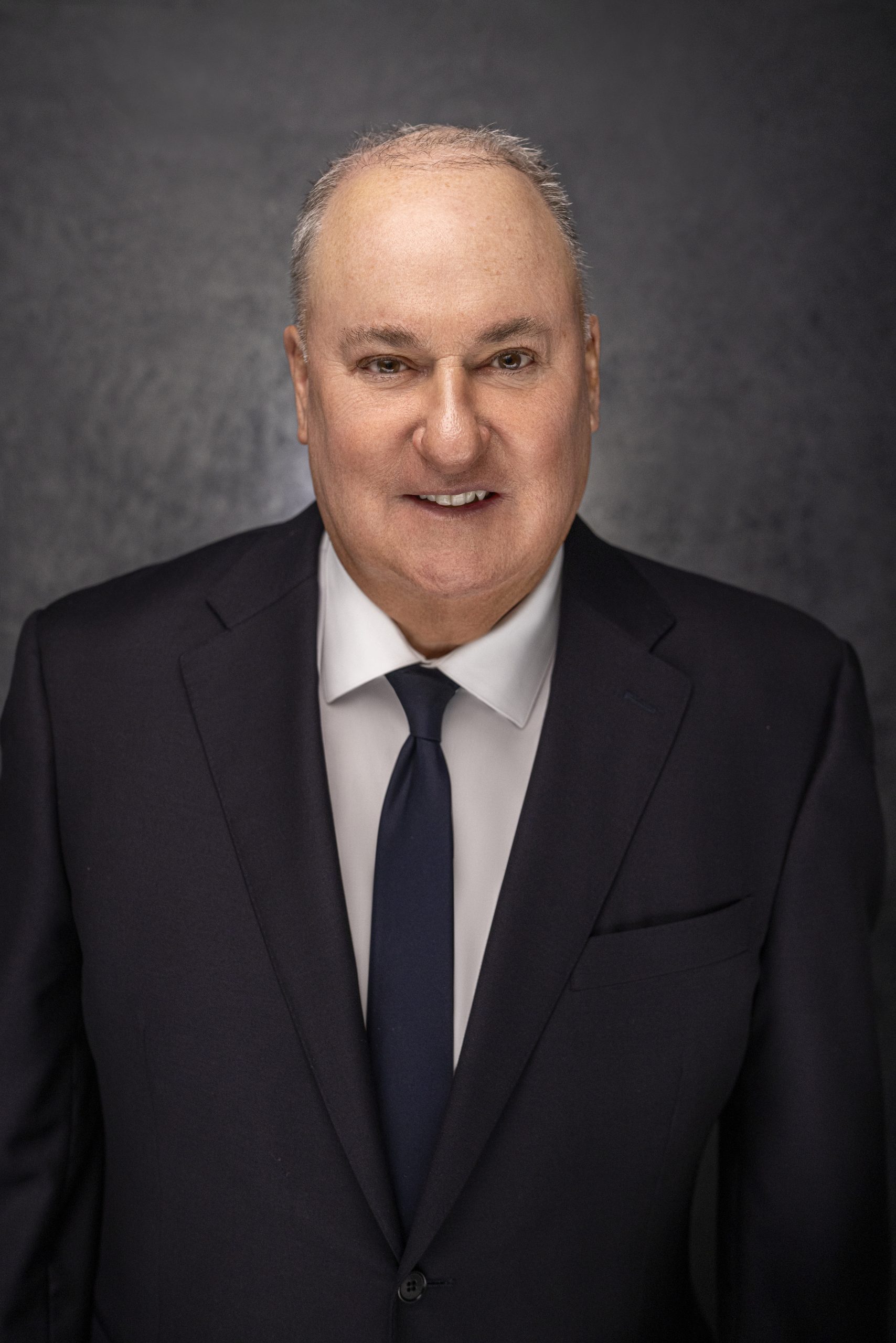 A middle-aged man with short gray hair, wearing a dark suit, white shirt, and navy tie, smiling slightly while standing against a dark, neutral background.