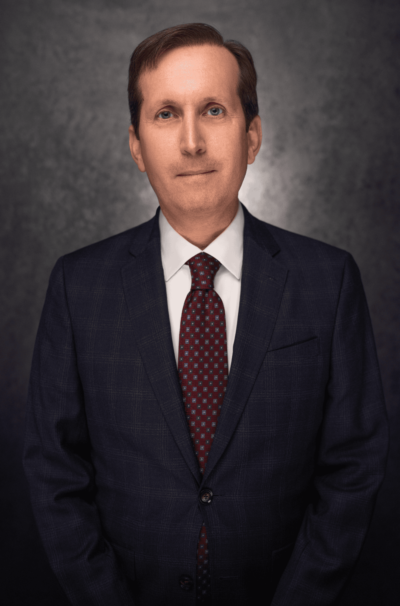 A middle-aged man with short brown hair, wearing a dark blue suit, white dress shirt, and patterned red tie, poses against a dark, textured background. He is facing the camera with a neutral expression.