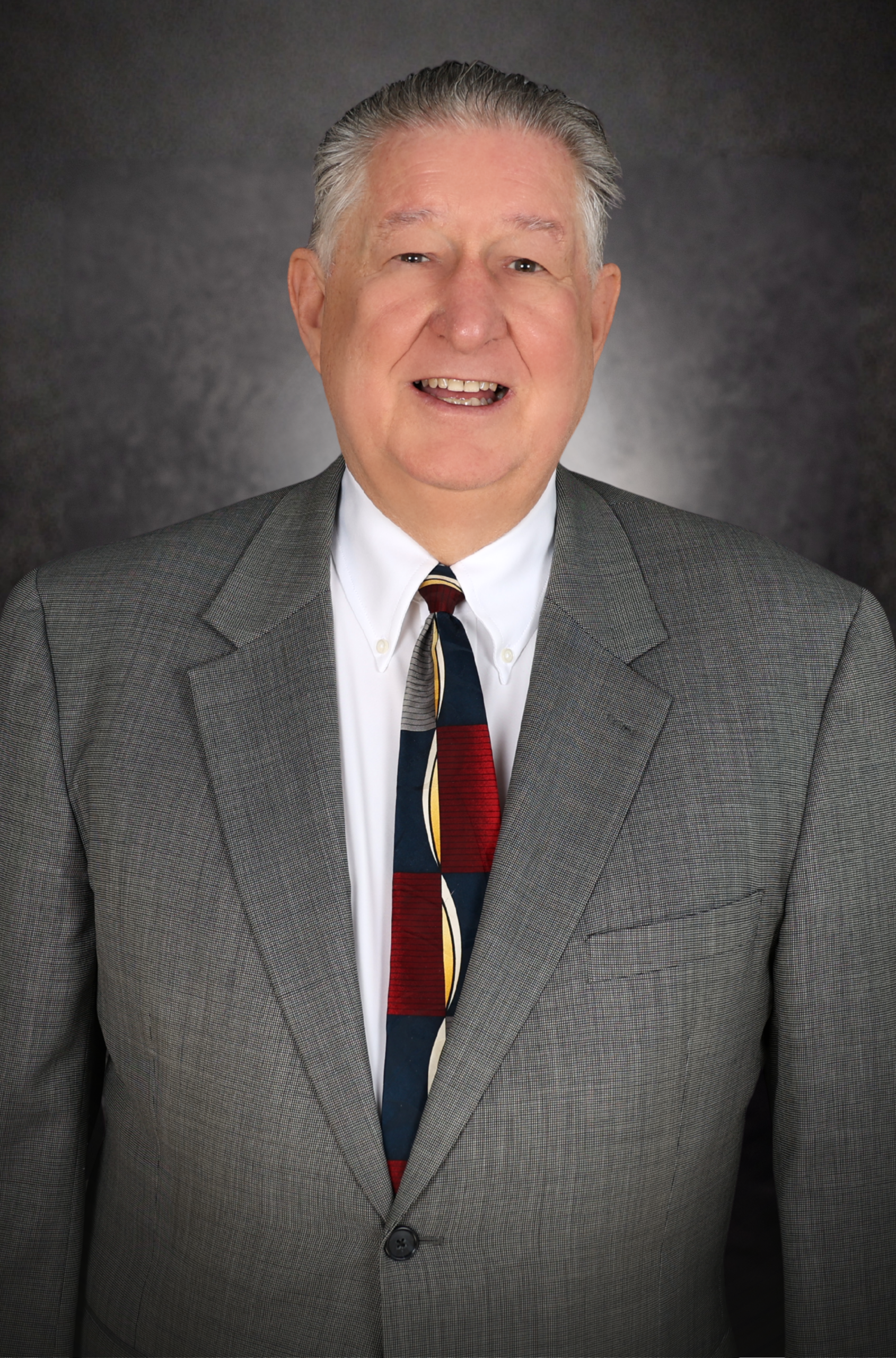 An older man with gray hair smiles while wearing a gray suit, white shirt, and a patterned tie, standing in front of a dark, textured background.