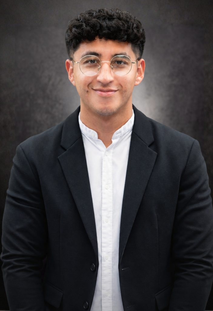 A young man wearing glasses, a white shirt, and a black blazer stands against a dark, textured background, smiling gently at the camera.