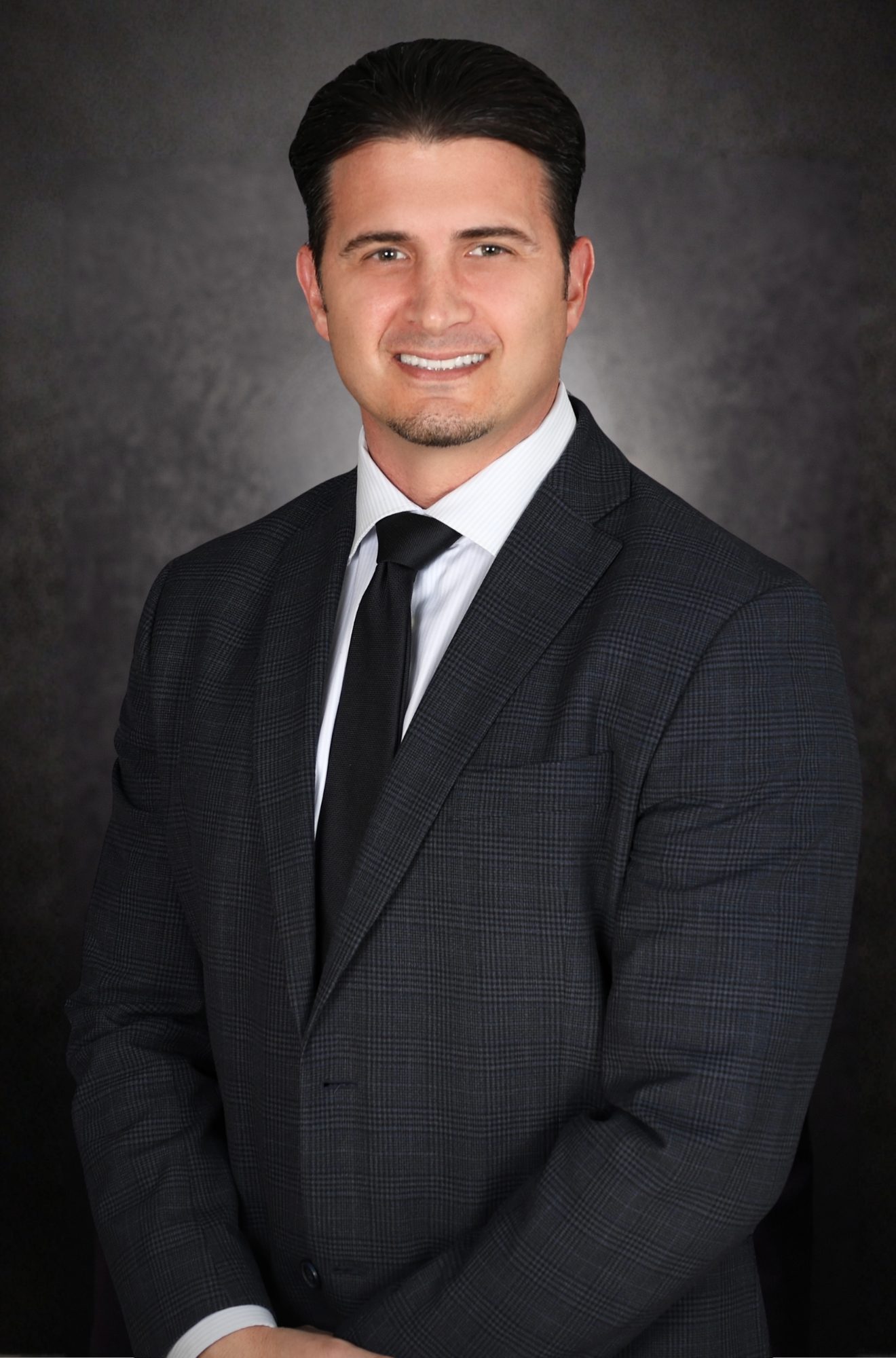 A man in a dark plaid suit, white shirt, and black tie stands smiling against a dark, textured background with his hands folded in front of him.