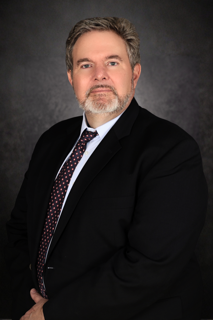 A middle-aged man with short gray hair and a beard, wearing a black suit, white shirt, and patterned tie, poses against a dark, textured background. He looks directly at the camera with a serious expression.