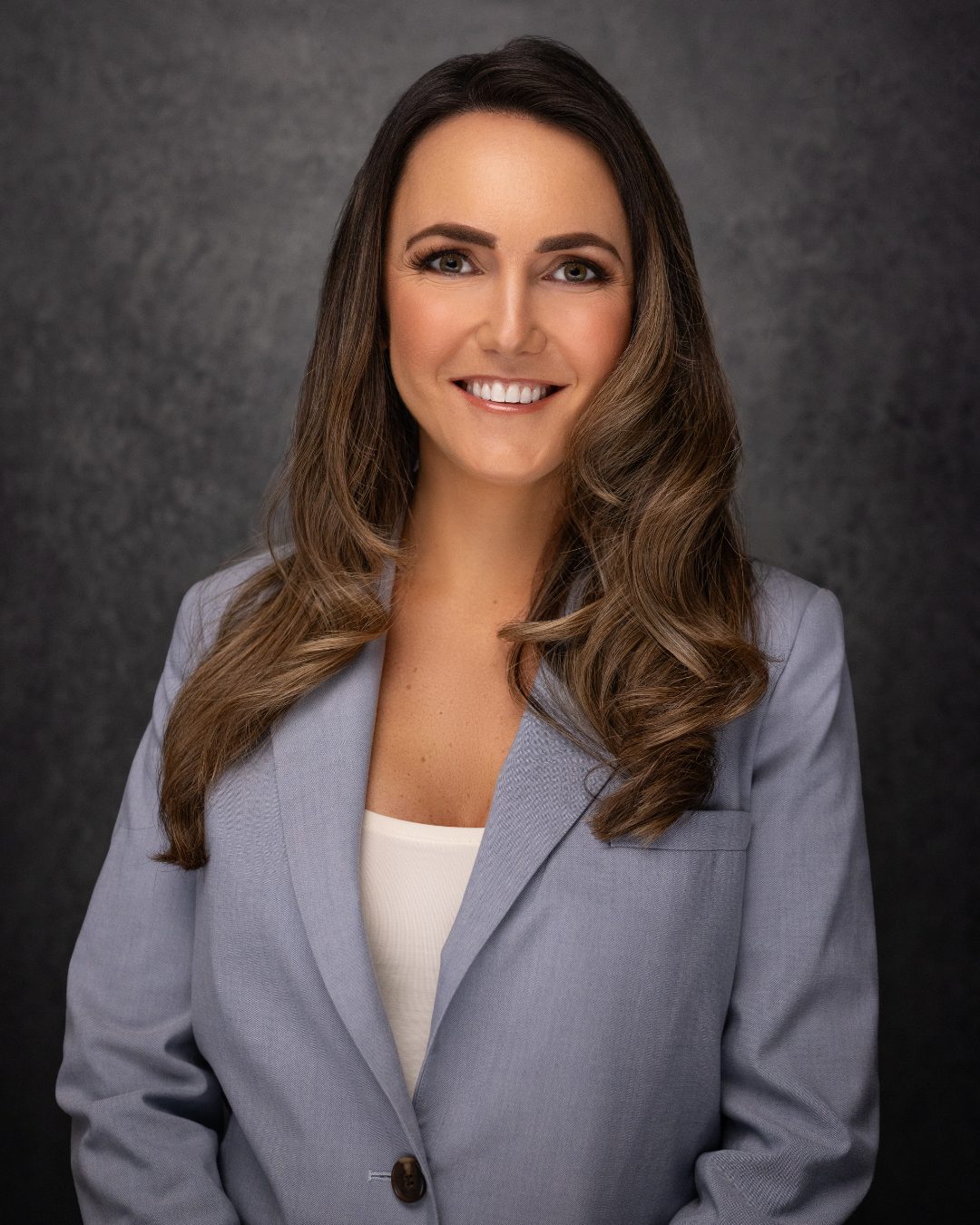 A woman with long brown hair wearing a light blue blazer and white top smiles at the camera against a dark textured background.