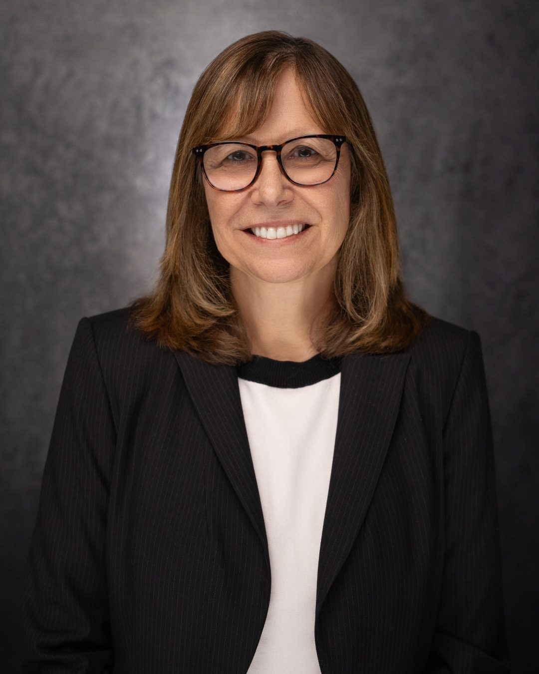 A woman with shoulder-length brown hair, glasses, and a black blazer over a white top smiles in front of a dark, neutral background.