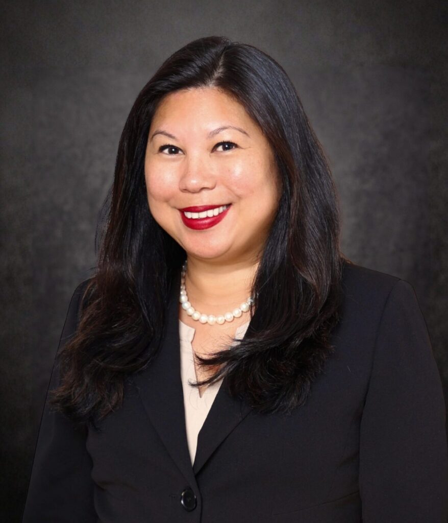 A woman with long dark hair, wearing a black blazer, beige top, pearl necklace, and red lipstick, smiles in front of a dark, textured background.
