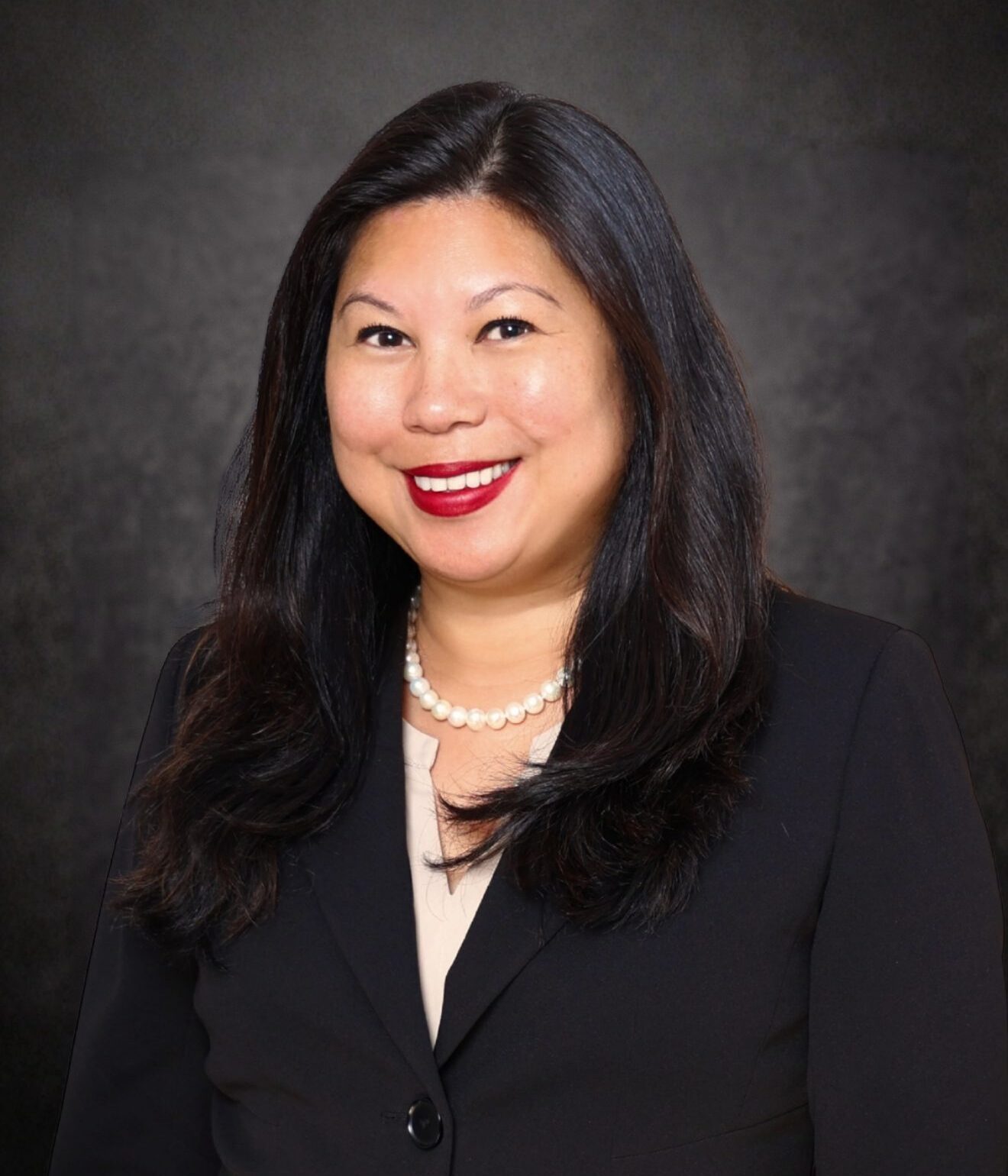 A woman with long dark hair, wearing a black blazer, beige top, pearl necklace, and red lipstick, smiles in front of a dark, textured background.
