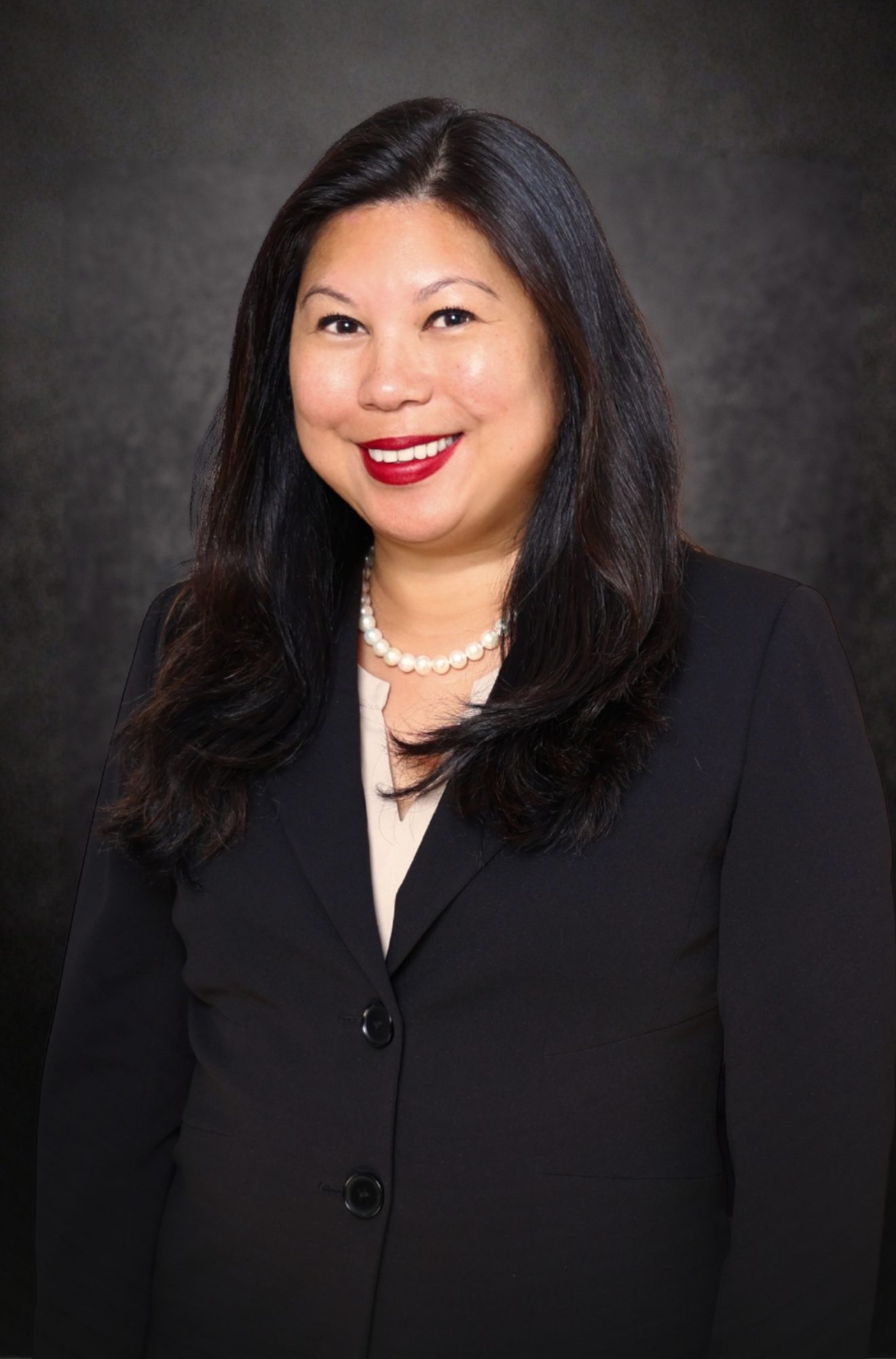 A woman with long dark hair, wearing a black blazer, beige top, pearl necklace, and red lipstick, smiles in front of a dark, textured background.